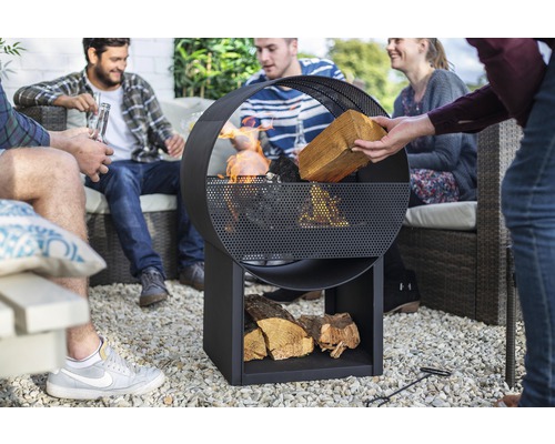 Skupina ľudí sedí v záhrade okolo kovového ohniska s úložným priestorom na drevo.