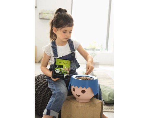 Dievča plní kvetináč v tvare tváre semenami trávy, drží obal so semenami, bio pečať