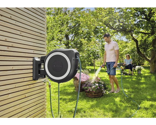 Box na záhradnú hadicu namontovaný na drevenej stene s mužom zalievajúcim kvety.