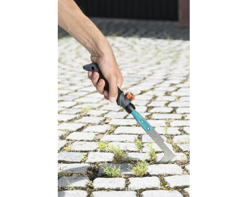 Ruka drží škrabku na škáry na odstraňovanie buriny medzi dlažobnými kockami.