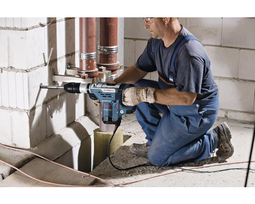 Remeselník vŕta vŕtacím kladivom otvor do steny, v pozadí vedú rúrky.