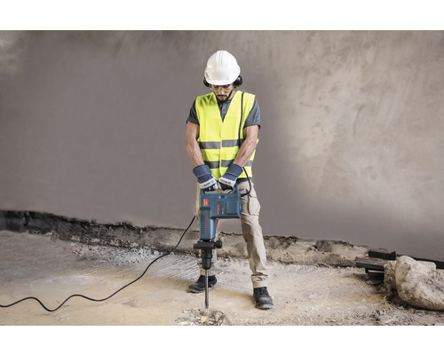 Stavebný robotník s ochrannou helmou, okuliarmi, ochranou sluchu, bezpečnostnou vestou a rukavicami obsluhuje zbíjacie kladivo.