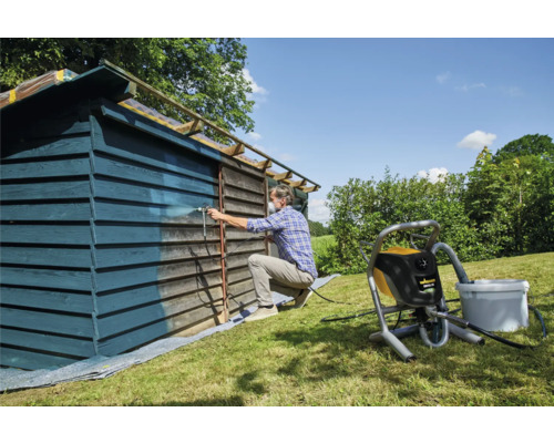 Muž strieka farbou drevenú búdu pomocou striekacieho systému