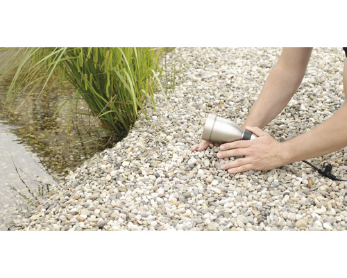 Muž inštaluje záhradný reflektor na štrkovej ceste vedľa jazierka s rastlinami.