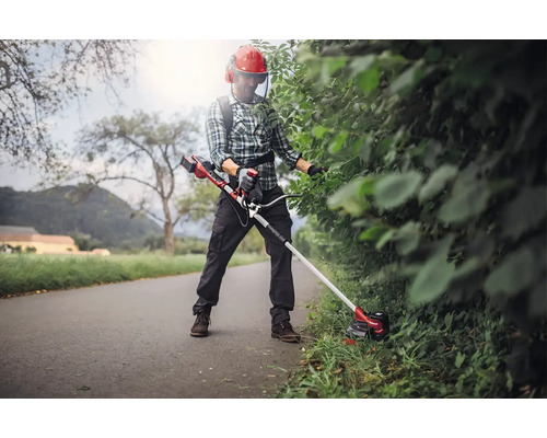 Osoba zastriháva živý plot pomocou akumulátorových nožníc na trávu, ktoré nosí s popruhom. Osoba má na sebe ochranný odev.