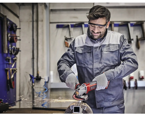 Muž nosí ochranné okuliare a pracuje s akumulátorovou uhlovou brúskou na kuse kovu v dielni.