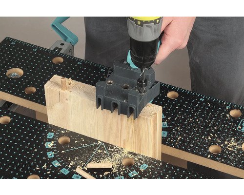 Osoba vŕta vŕtačkou otvor do kusa dreva pomocou prípravku na kolíkovanie na pracovnom stole.