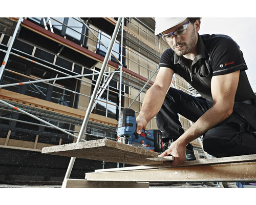 Stavebný robotník s helmou a ochrannými okuliarmi reže priamočiarou pílou drevenú dosku na stavenisku. V pozadí je vidieť lešenie. Logo Bosch.