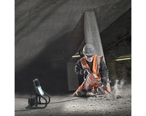 Stavebný robotník používa zbíjacie kladivo na stavenisku pod betónovou konštrukciou, osvetlené pracovným svetlom.