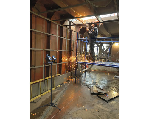 Muž na lešení zvára kov zváračkou v priemyselnom prostredí.