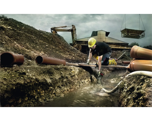 Stavebný robotník pracuje v priekope s rúrkami, bagrom a žeriavom v pozadí.