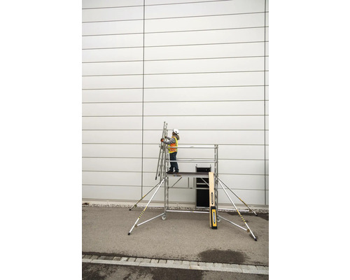 Osoba s ochrannou prilbou a bezpečnostnou vestou stojí na pojazdnom lešení a drží rebrík pri stene.