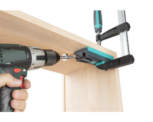 Akumulátorový skrutkovač vŕta s vŕtacím puzdrom drevenú dosku, ktorá je upevnená skrutkovou svorkou.