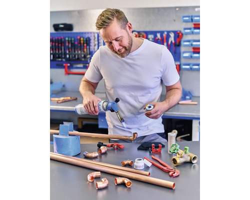 Muž spájkuje medené rúry plynovým horákom, obklopený medenými rúrkovými tvarovkami a náradím