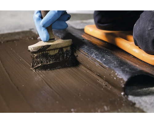 Osoba v modrých rukaviciach nanáša štetcom hydroizolačnú suspenziu.