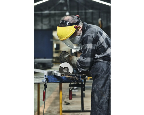 Remeselník s ochranným štítom a rukavicami reže kov brúskou na zveráku.