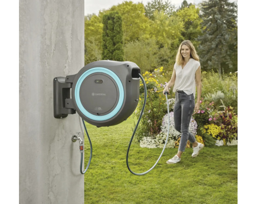 Záhradná scéna s boxom na hadicu na zavlažovanie záhrady, namontovaným na stene. Žena drží koniec hadice.