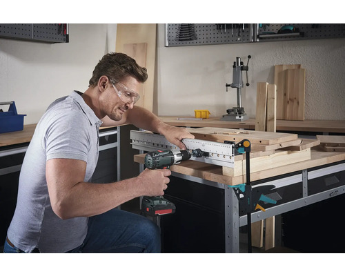 Muž vŕta vŕtačkou cez drevenú dosku s vŕtacou šablónou na pracovnom stole.