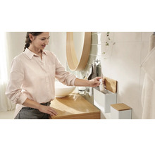 Svetlá kúpeľňa s umývadlom, zrkadlom a ženou, ktorá berie fľaštičku z nástenného regálu.