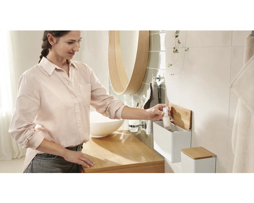 Svetlá kúpeľňa s umývadlom, zrkadlom a ženou, ktorá berie fľaštičku z nástenného regálu.