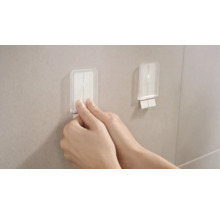 Dva priehľadné samolepiace háčiky sú pripevnené na stene. Jedna ruka ťahá za jeden z háčikov.