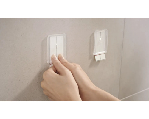 Dva priehľadné samolepiace háčiky sú pripevnené na stene. Jedna ruka ťahá za jeden z háčikov.