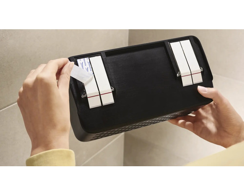 Osoba pripevňuje čiernu nástennú policu lepiacimi prúžkami na stenu