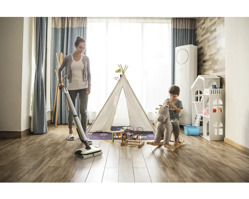 Žena umýva drevenú podlahu v detskej izbe pomocou akumulátorového mopu, zatiaľ čo sa dieťa hrá na hojdacom koni.