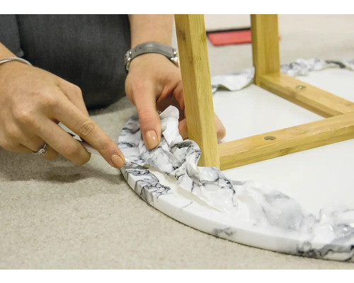 Detailný záber osoby aplikujúcej fóliu s mramorovým vzorom na odkladací stolík s drevenými nohami