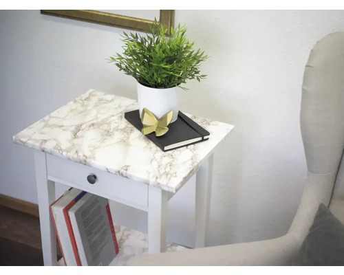 Dekoratívne aranžmán na bielom odkladacom stolíku s mramorovým vzhľadom, pozostávajúce z rastliny v kvetináči, zápisníka a ozdobnej figúrky.