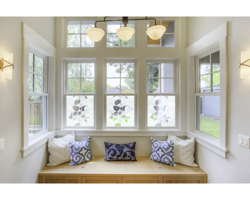 Svetlý arkier s oknami zdobenými rastlinnými motívmi na okenné fólie, lavica s vankúšmi, stropné svietidlo a nástenné lampy