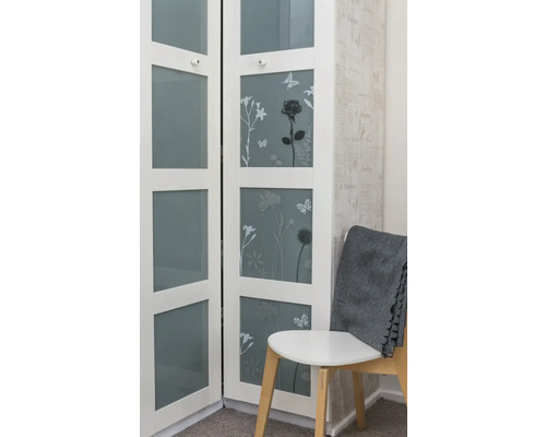 Biela skriňa s kvetinovými motívmi a sklenenými dverami vedľa stoličky so sivou dekou