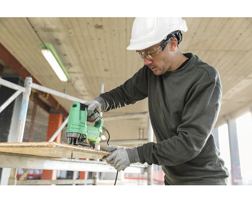 Muž reže drevenú dosku priamočiarou pílou značky Hikoki a má na sebe ochranné oblečenie.