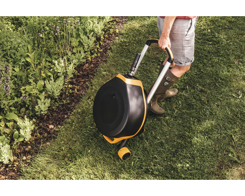 Záhradná scéna s rotačným kompostérom na kolieskach, ktorý drží osoba na trávniku