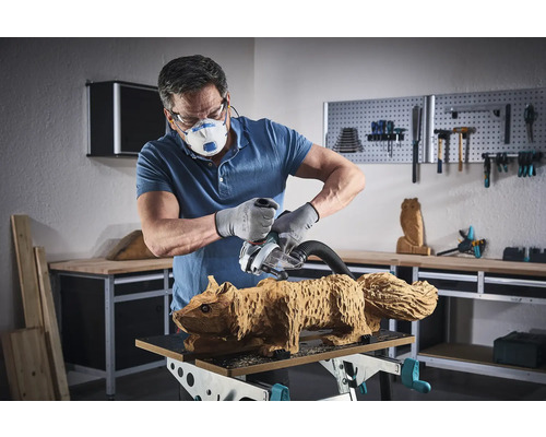 Muž brúsi brúsnym strojom drevenú figúrku veveričky. Má na sebe masku, ochranné okuliare a rukavice.