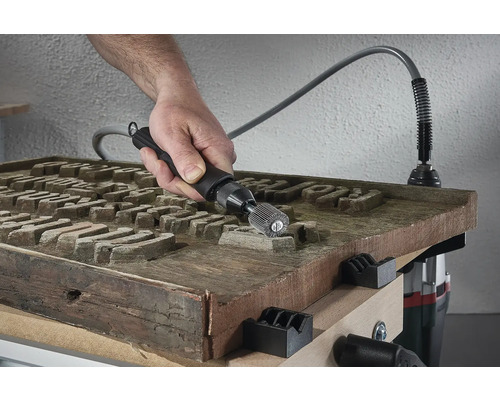 Ruka s brúsnym nástrojom opracováva drevenú dosku s rytinou na pracovnom stole