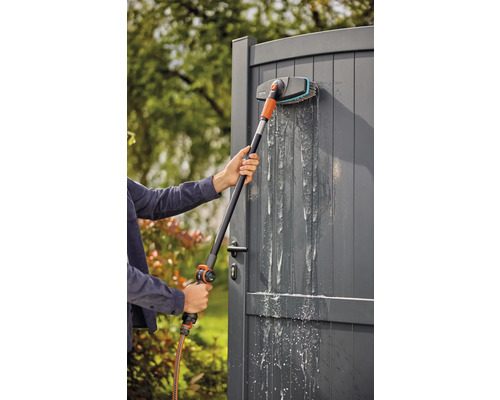 Osoba čistí sivú záhradnú bránu umývacou kefou a záhradnou hadicou.