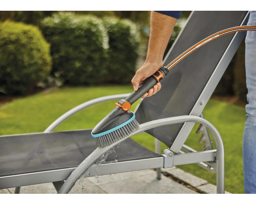 Osoba čistí záhradné ležadlo kefou a vodnou hadicou.