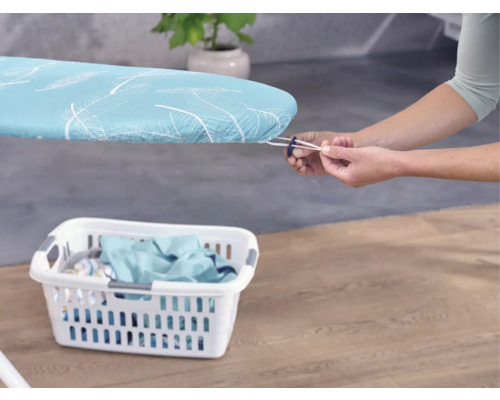 Osoba upevňuje poťah na žehliacu dosku s gumičkou, v popredí kôš na bielizeň