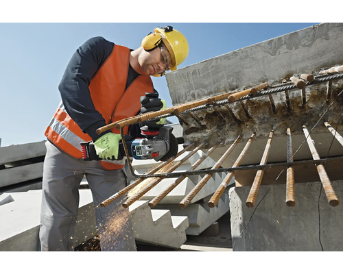 Stavebný robotník s ochrannou vestou, ochrannými okuliarmi, chráničmi sluchu a helmou oddeľuje armovacie železo uhlovou brúskou.