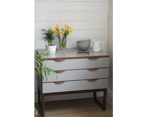 Komoda s tromi zásuvkami, na ktorej je rastlina, narcisy vo váze, misky, okuliare a džbán.