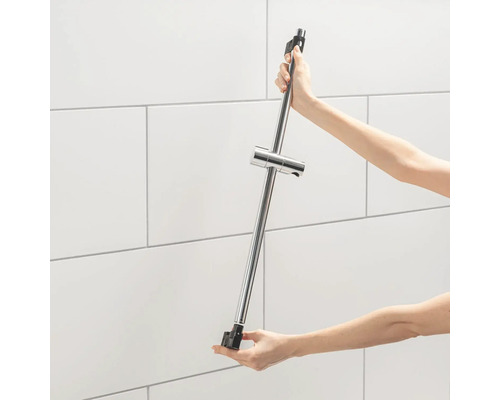 Osoba drží sprchovú tyč s držiakom sprchovej hlavice pred obloženou stenou.