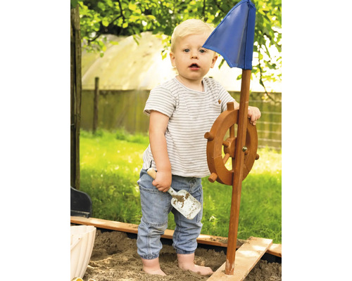 Batoľa sa hrá s hracou loďou s volantom a modrou vlajkou v pieskovisku