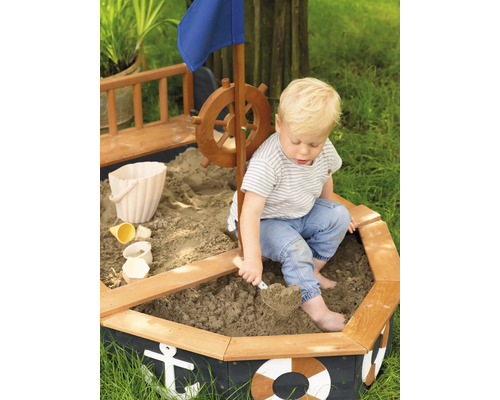 Dieťa sa hrá v pieskovisku v tvare lode s kormidlom, vlajkou a námorníckymi motívmi.