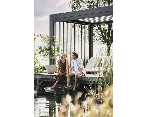 Pár sedí na drevenom móle pri vode pod pergolou. Záhradný dizajn s lounge nábytkom a výsadbou.