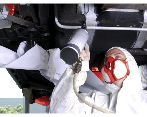 Osoba v ochrannom odeve nosí respirátor a strieka ochranu podvozku na zdvihnuté auto.