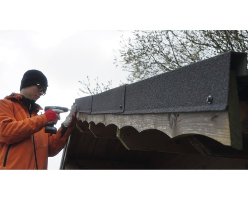 Muž pripevňuje lepenku akumulátorovým skrutkovačom na drevenú kôlňu.