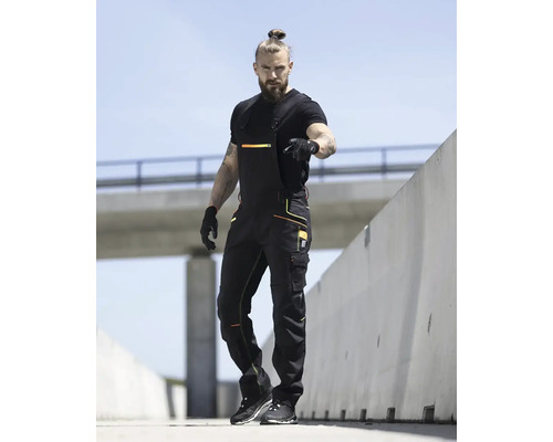 Muž v čiernom pracovnom oblečení s náprsenkou pred betónovou stenou