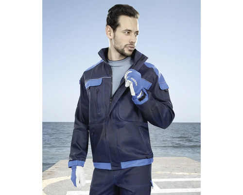 Muž má na sebe tmavomodrú pracovnú bundu s modrými akcentmi a pracovné rukavice na pozadí mora.