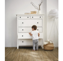 Dieťa stojí pred bielou komodou so zásuvkami a okrúhlymi úchytkami vo svetlej miestnosti.